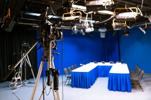 A full-production studio is shown from the view of behind the camera. Multiple lights and cables are mounted to the ceiling above 3 tables.